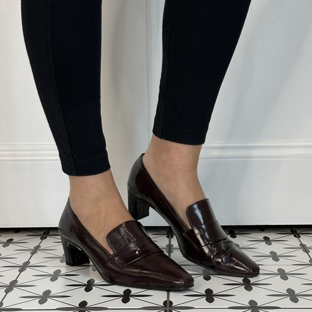 A pair of Unisa dark brown patent leather slip-on pumps with kitten heels, shown on a model wearing black leggings on a black and white patterned tile floor.