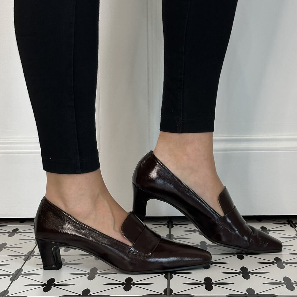 A pair of Unisa dark brown patent leather slip-on pumps with kitten heels, shown on a model wearing black leggings on a black and white patterned tile floor.