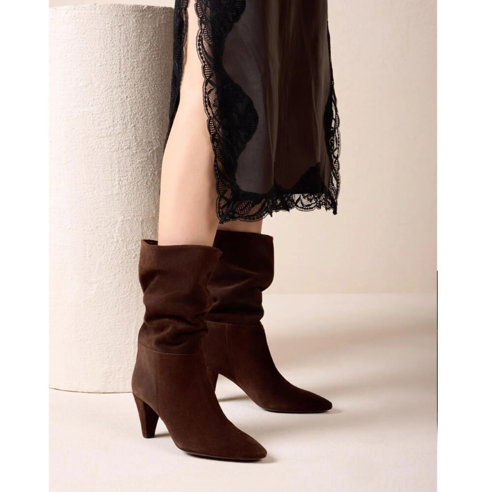 A pair of Unisa dark brown suede mid-calf boots with a slouchy shaft and low block heel, shown on a model wearing a sheer black lace-trimmed skirt against a neutral beige studio background.
