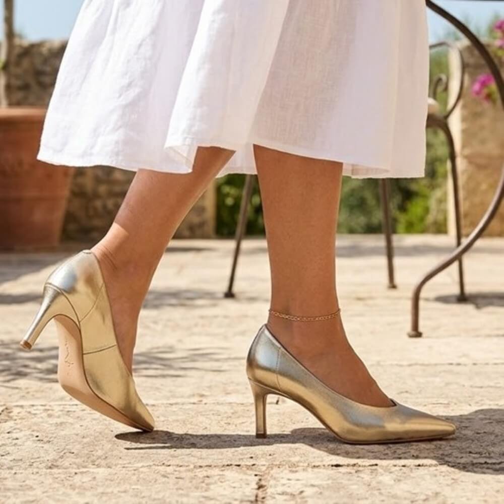 Unisa Lenis Gold high-heeled shoes worn with a white skirt on a stone pavement.
