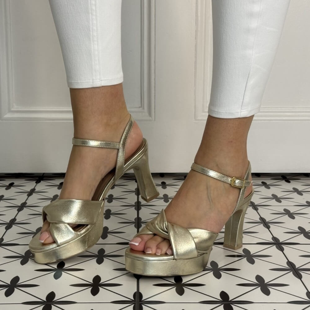 Gold high-heeled sandals worn with white pants on a patterned floor.