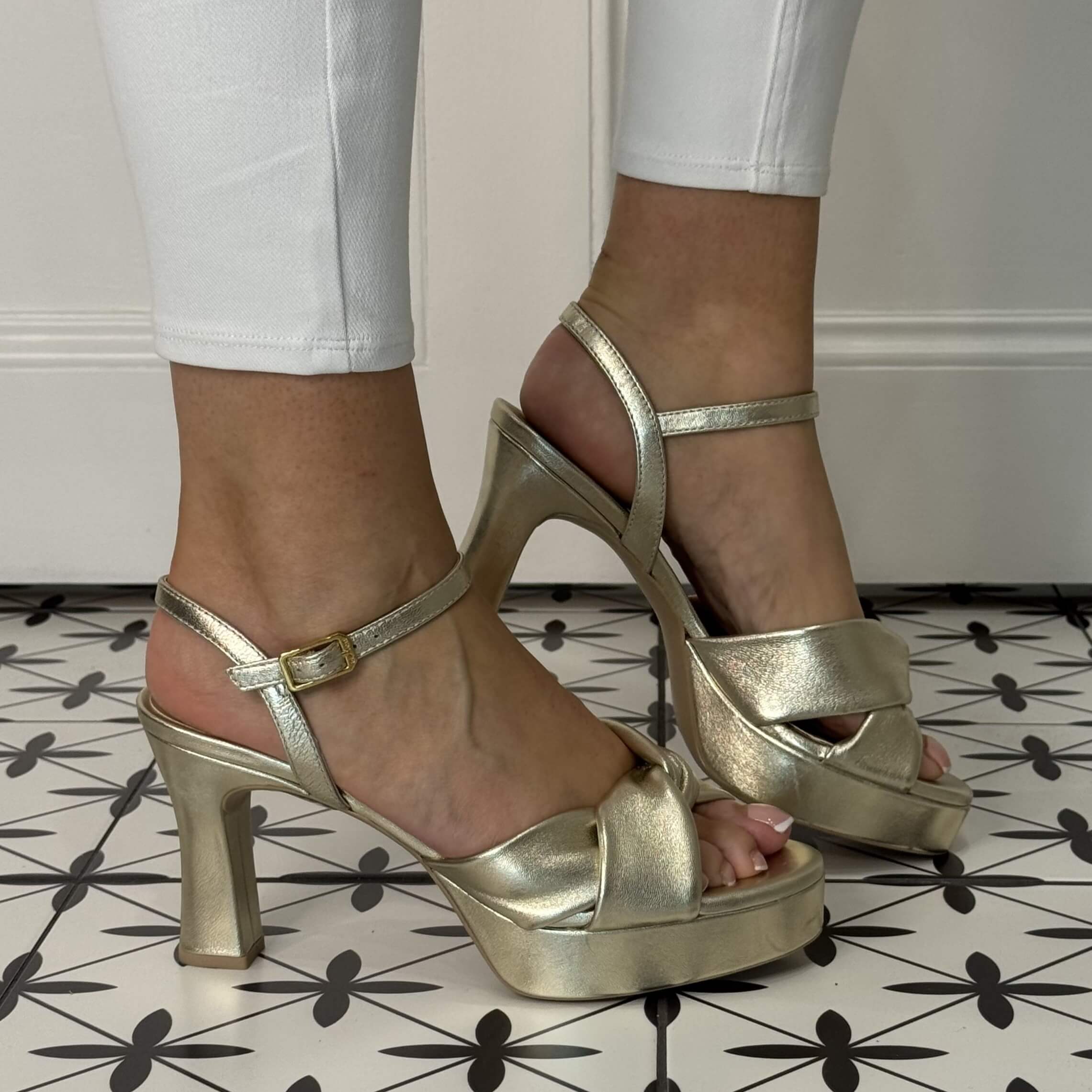 Gold high-heeled sandals worn with white pants on a patterned floor.