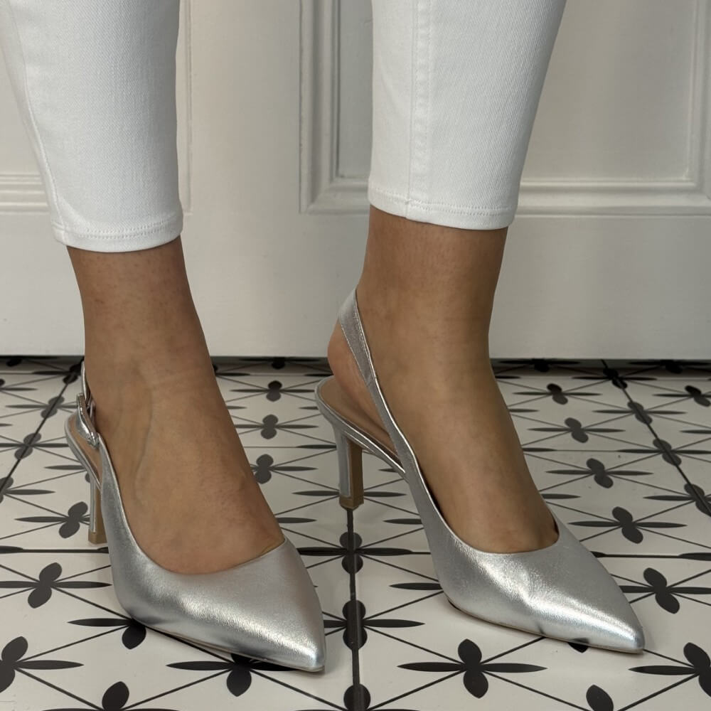 Silver high-heeled shoes worn with white pants on a patterned floor. unisa lanz