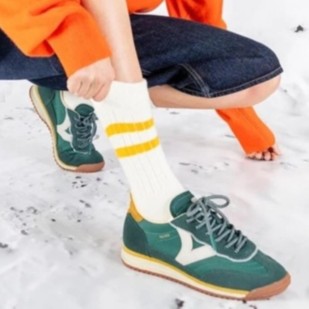 Green sneakers with brown soles worn by a person sitting on a snowy ground. Green, white and yellow sneaker with a white logo on a white background Victoria Saturino Sneakers 1158101