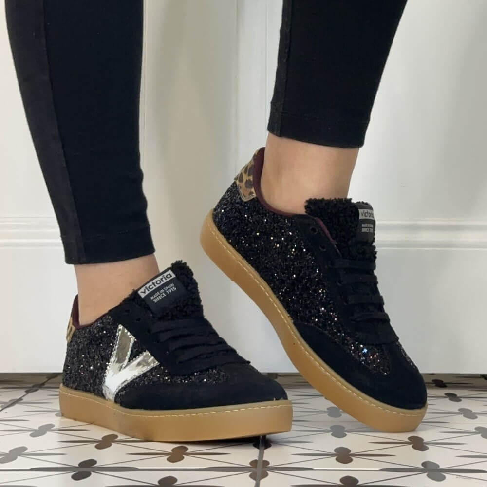 A person wearing Victoria black sneakers covered in black glitter, with a metallic silver V-shaped side stripe, leopard-print heel tabs, and tan gum soles. The person is wearing black leggings, standing on a tiled floor.