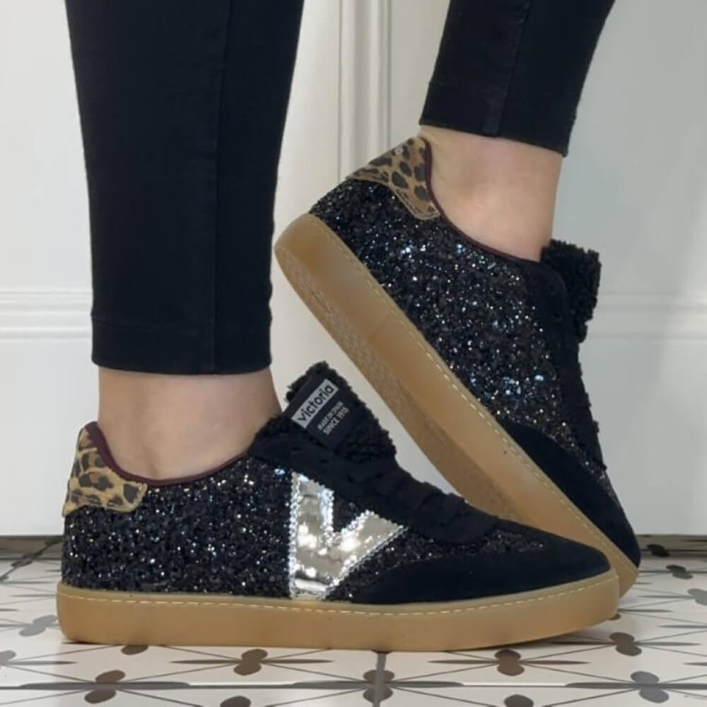 A person wearing Victoria black sneakers covered in black glitter, with a metallic silver V-shaped side stripe, leopard-print heel tabs, and tan gum soles. The person is wearing black leggings, standing on a tiled floor.