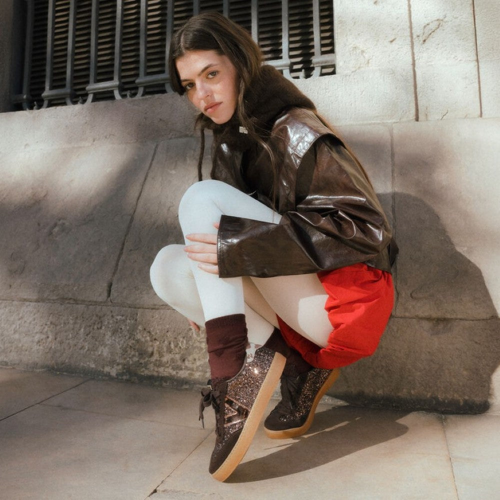 A model crouches against a stone wall wearing a shiny dark brown jacket, white tights, red shorts, and Victoria brown sparkle lace-up shoes with tan soles.