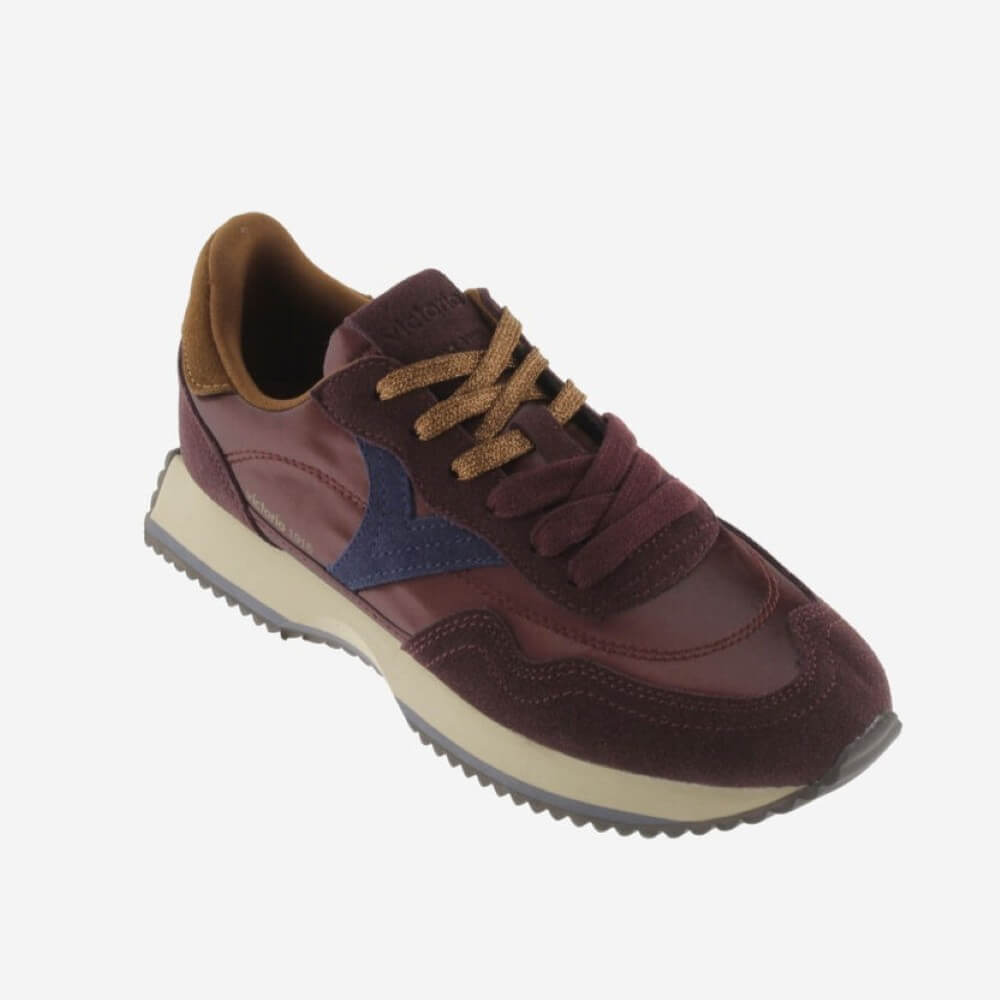 A single low-top sneaker shown at a three-quarter angle on a light gray background. The shoe features a color-blocked design with a burgundy upper, a navy blue chevron accent on the side, a tan collar lining, brown laces, and a thick cream midsole with a dark gray tread.
