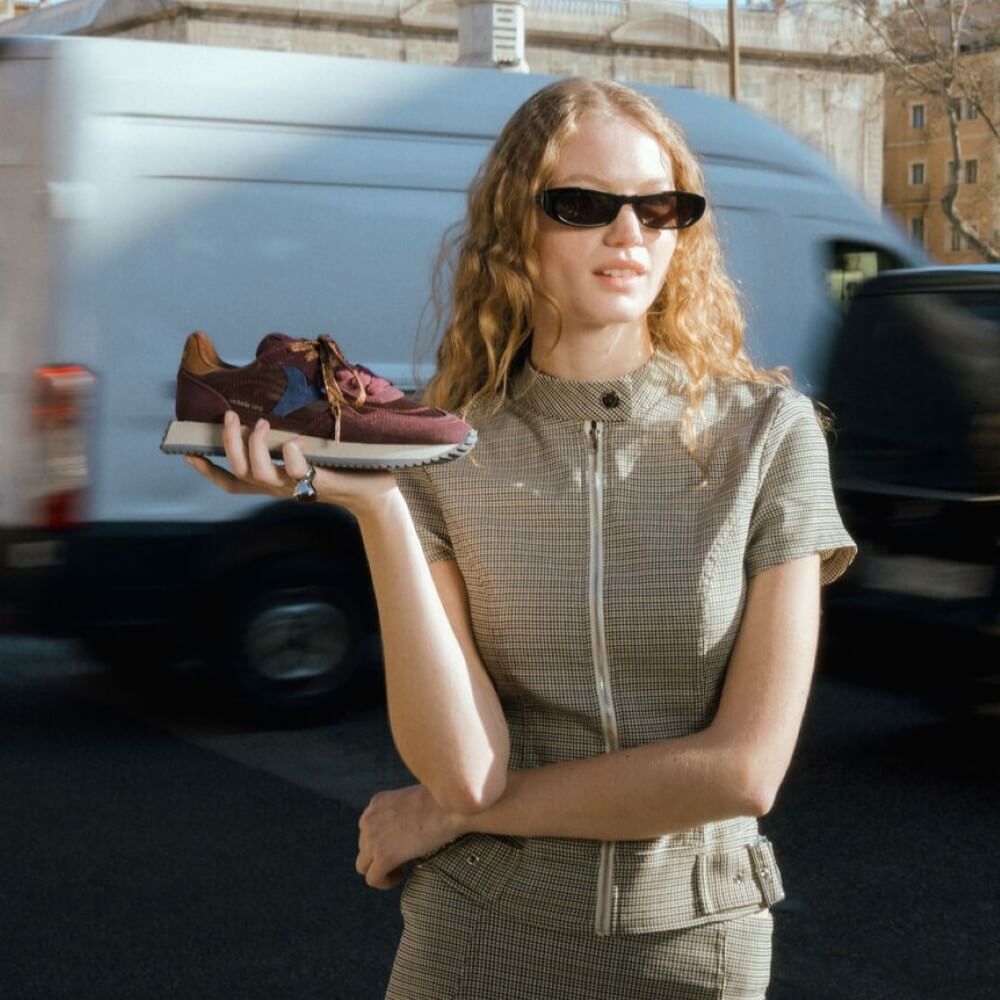 A woman wearing sunglasses and a textured beige outfit holds up a Victoria brown suede sneaker with colourful star details on the side. The background shows a blurred street scene with a white van.