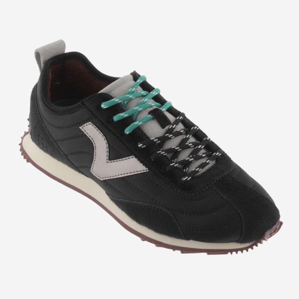A black Victoria sneaker with a metallic silver V logo on the side, teal laces, and a cream midsole, shown against a light grey background.