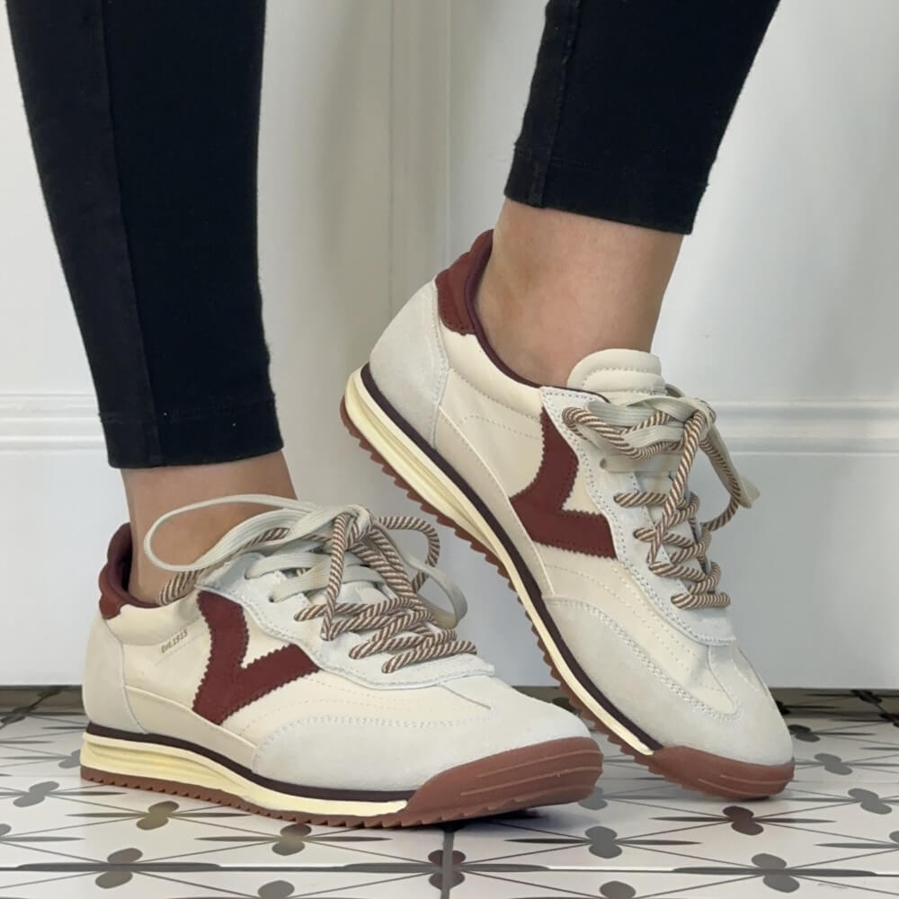 A pair of Victoria women's retro-style sneakers shown being worn, with black leggings visible on the lower legs. The shoes are off-white with dark burgundy side panels and heel tabs, cream midsoles, and beige rope laces.
