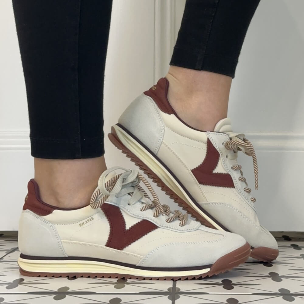 A pair of Victoria women's retro-style sneakers shown being worn, with black leggings visible on the lower legs. The shoes are off-white with dark burgundy side panels and heel tabs, cream midsoles, and beige rope laces.