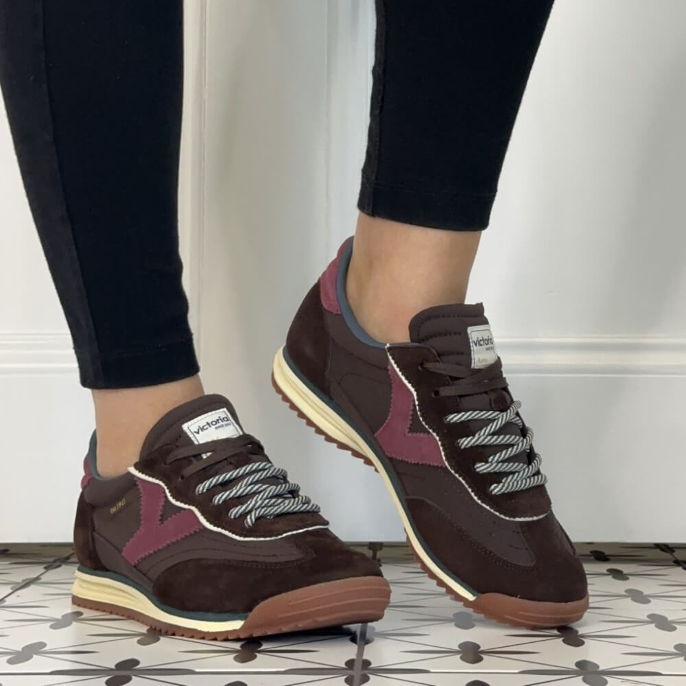 A person wearing dark brown and burgundy Victoria sneakers with cream midsoles, shown from the calves down against a white wall and patterned tile floor.