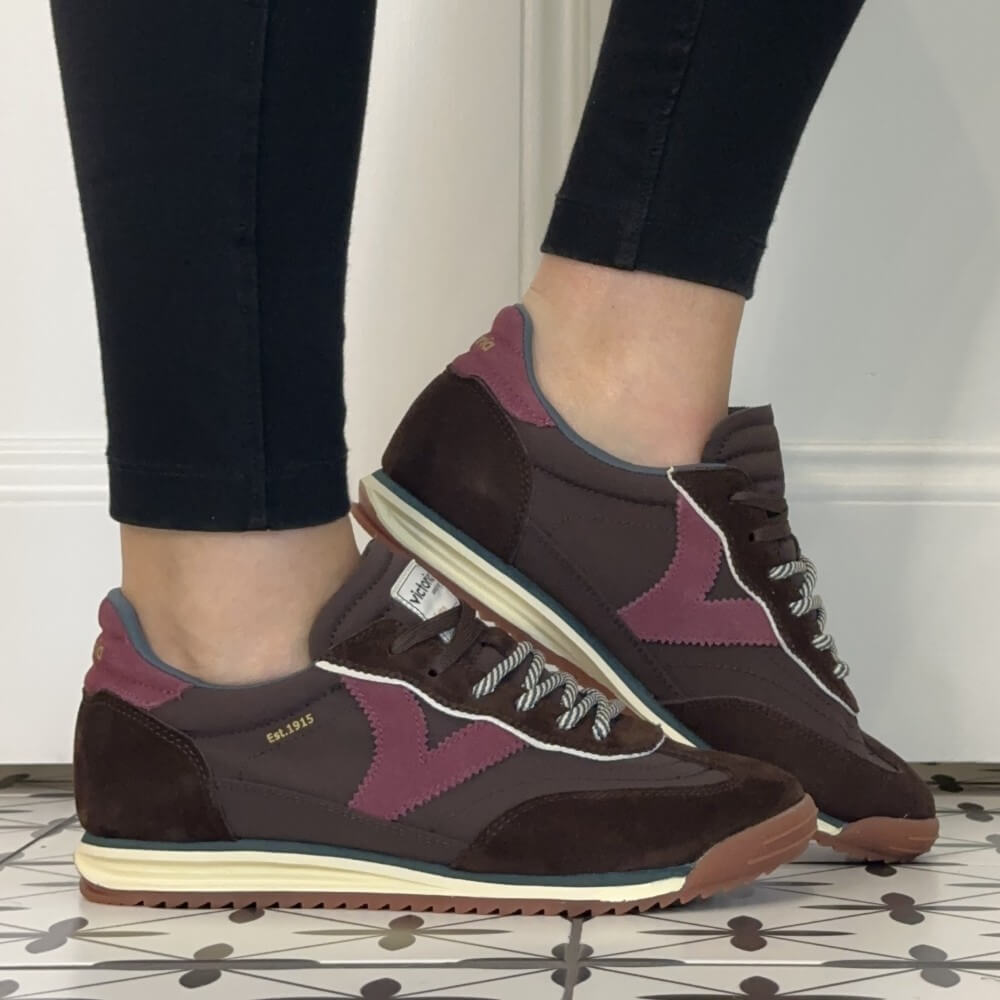 A person wearing dark brown and burgundy Victoria sneakers with cream midsoles, shown from the calves down against a white wall and patterned tile floor.