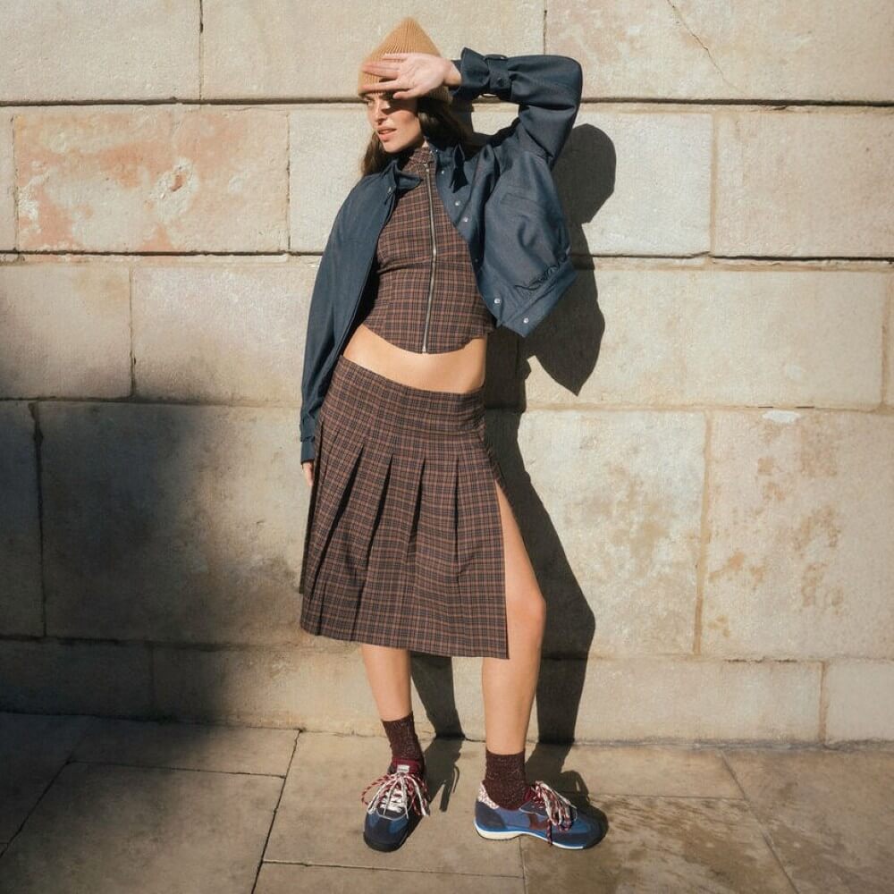 A model wearing a brown and black plaid crop top and matching pleated skirt, styled with a dark blue bomber jacket, brown socks, and blue Victoria sneakers, posing against a stone wall.