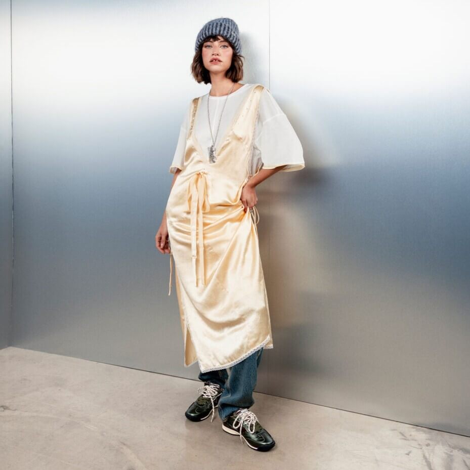 A model wearing a cream satin wrap dress over a white t-shirt, blue jeans, and Victoria Khaki Green sneakers, standing against a metallic silver wall.