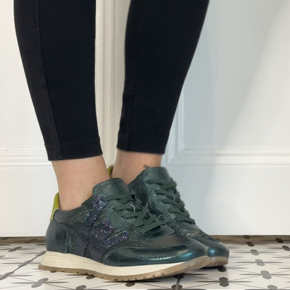 A person wearing Wonders dark green textured leather sneakers with a glittery purple side stripe, paired with black leggings, standing on a patterned tile floor against a white wall.