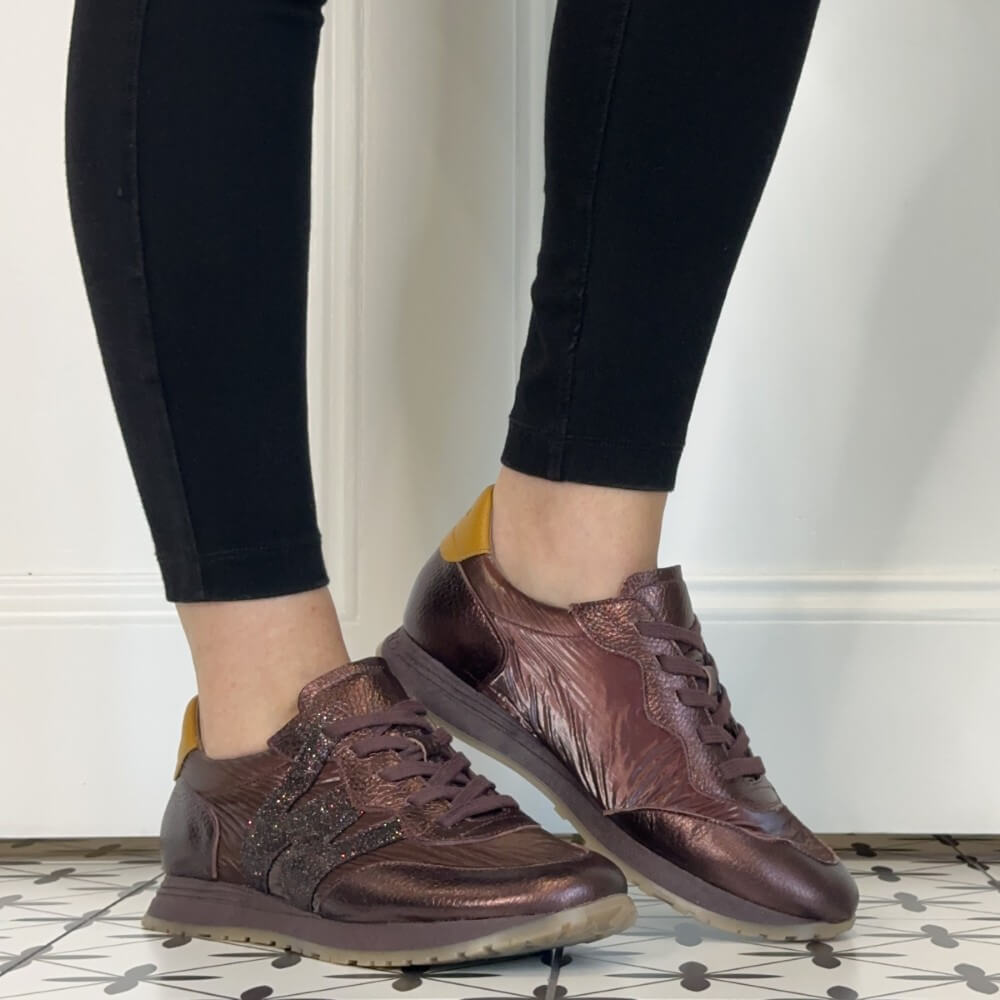 A person wearing black leggings and Wonders wine textured sneakers with yellow heel accents, standing on a patterned tile floor against a white wall.