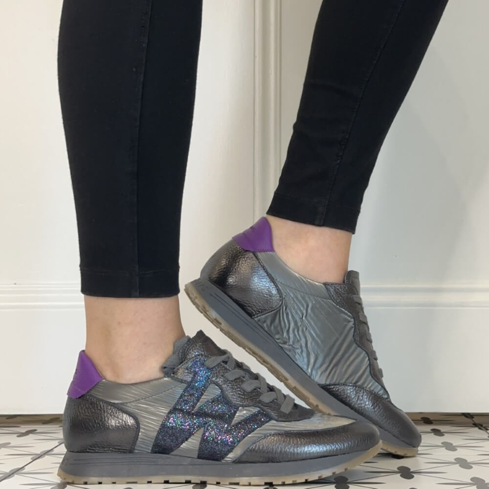 A person wearing Wonders sneakers in a home setting. The sneakers are metallic grey with a textured leather upper, a glittery navy side stripe, and a purple heel tab.