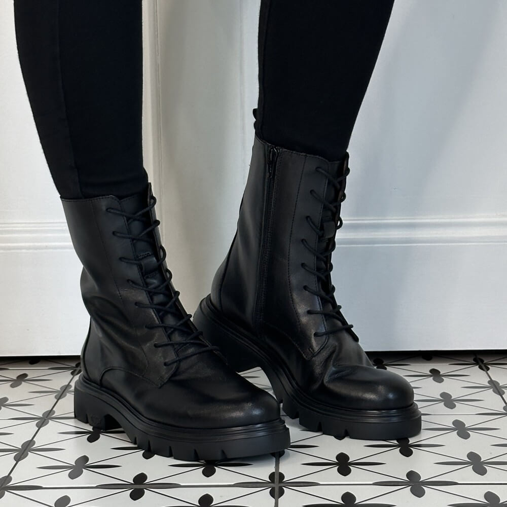 A pair of Wonders black leather lace-up combat boots with side zippers and chunky lug soles, worn with black leggings on a black-and-white patterned tile floor.