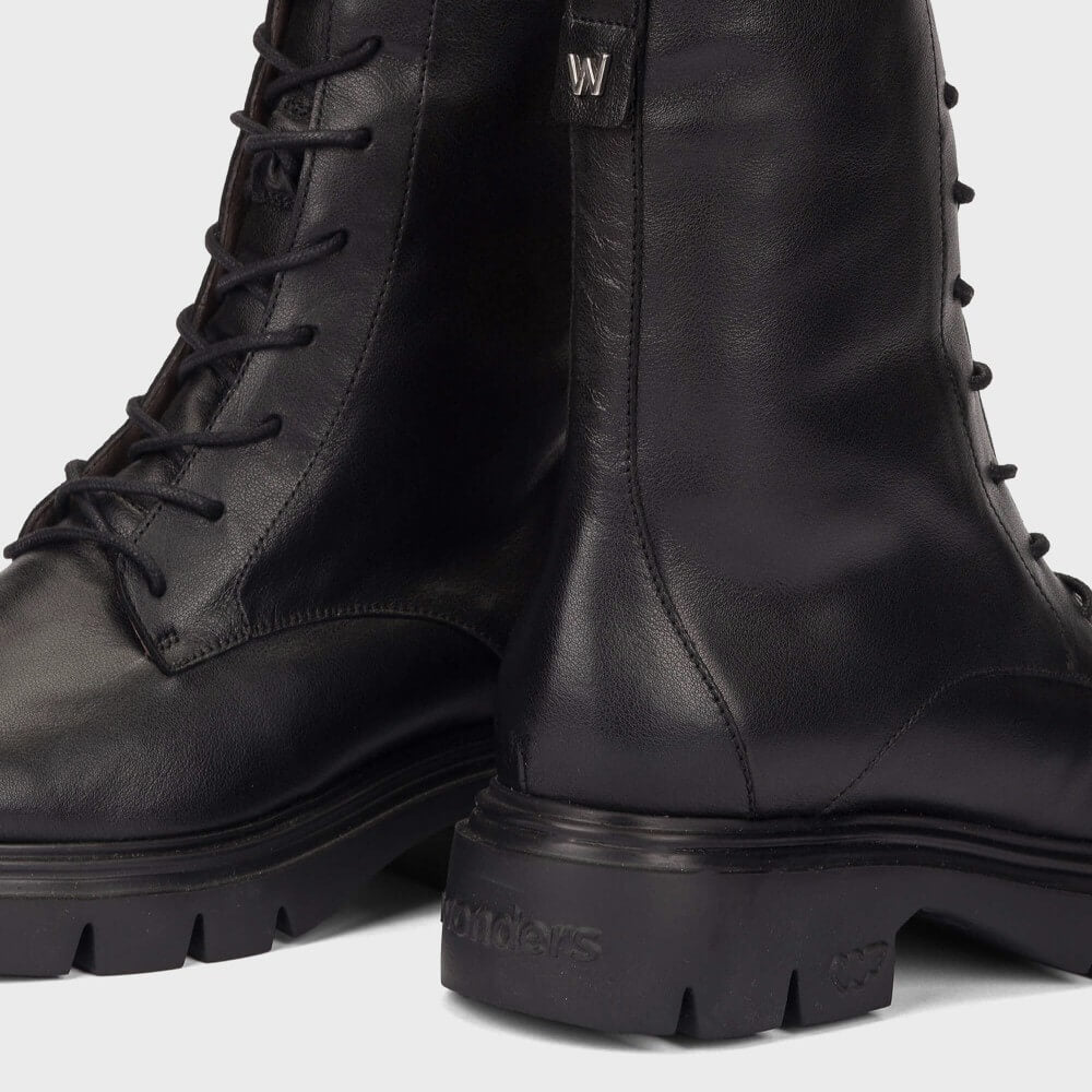 A close-up studio shot of a pair of black leather lace-up boots with chunky lug soles, showing the side profile and heel details against a light gray background.