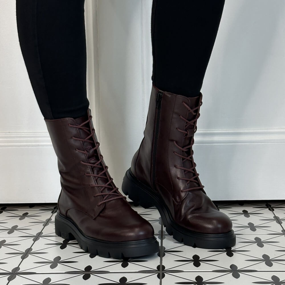 A pair of Wonders Wine leather lace-up combat boots with side zippers and chunky black soles, shown on a person wearing black leggings standing on a black-and-white patterned tile floor.
