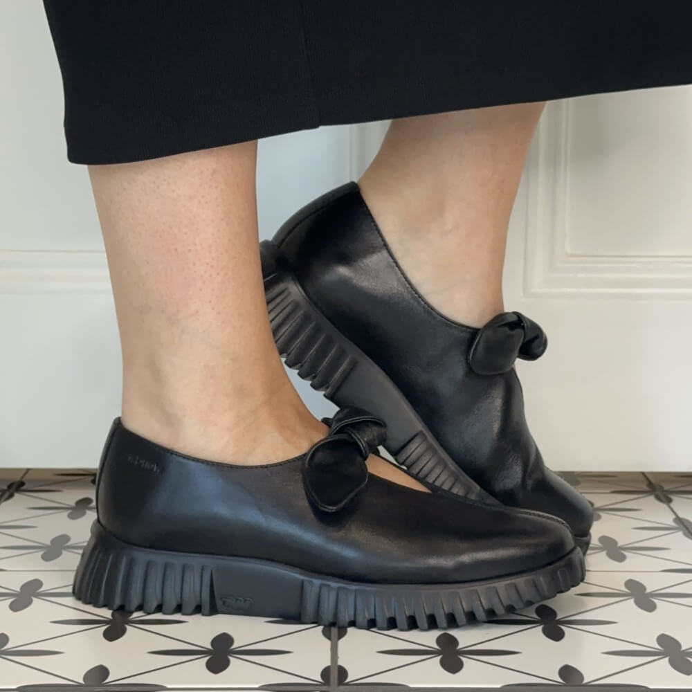 Wonders Black leather Mary Jane loafers with chunky lug soles and bow details, shown on a person's feet standing on a patterned tile floor.