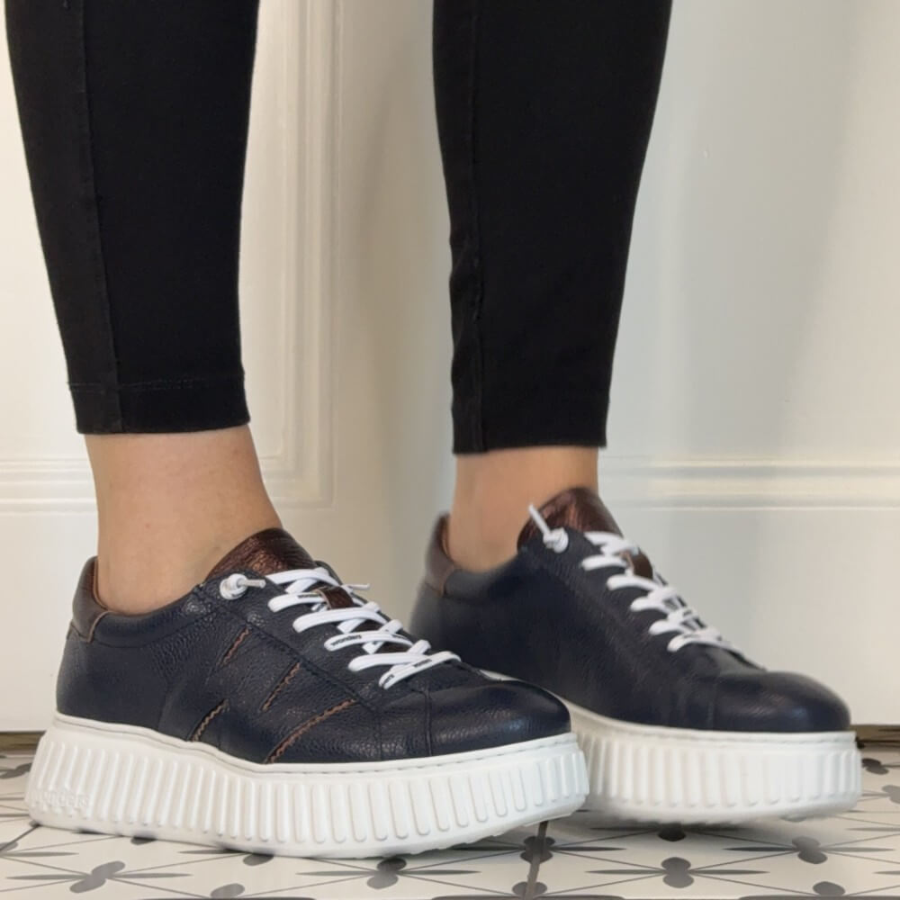 A person wearing Wonders Navy platform sneakers with white laces and thick white platform soles, paired with black leggings, standing on a patterned tile floor against a white panelled wall.