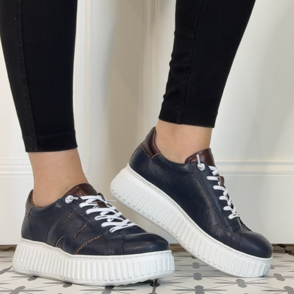 A person wearing Wonders Navy platform sneakers with white laces and thick white platform soles, paired with black leggings, standing on a patterned tile floor against a white panelled wall.