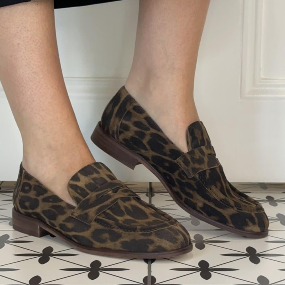 A pair of Wonders brown and black leopard print penny loafers worn on a person's feet, standing on a black and white patterned tile floor against a white paneled wall.