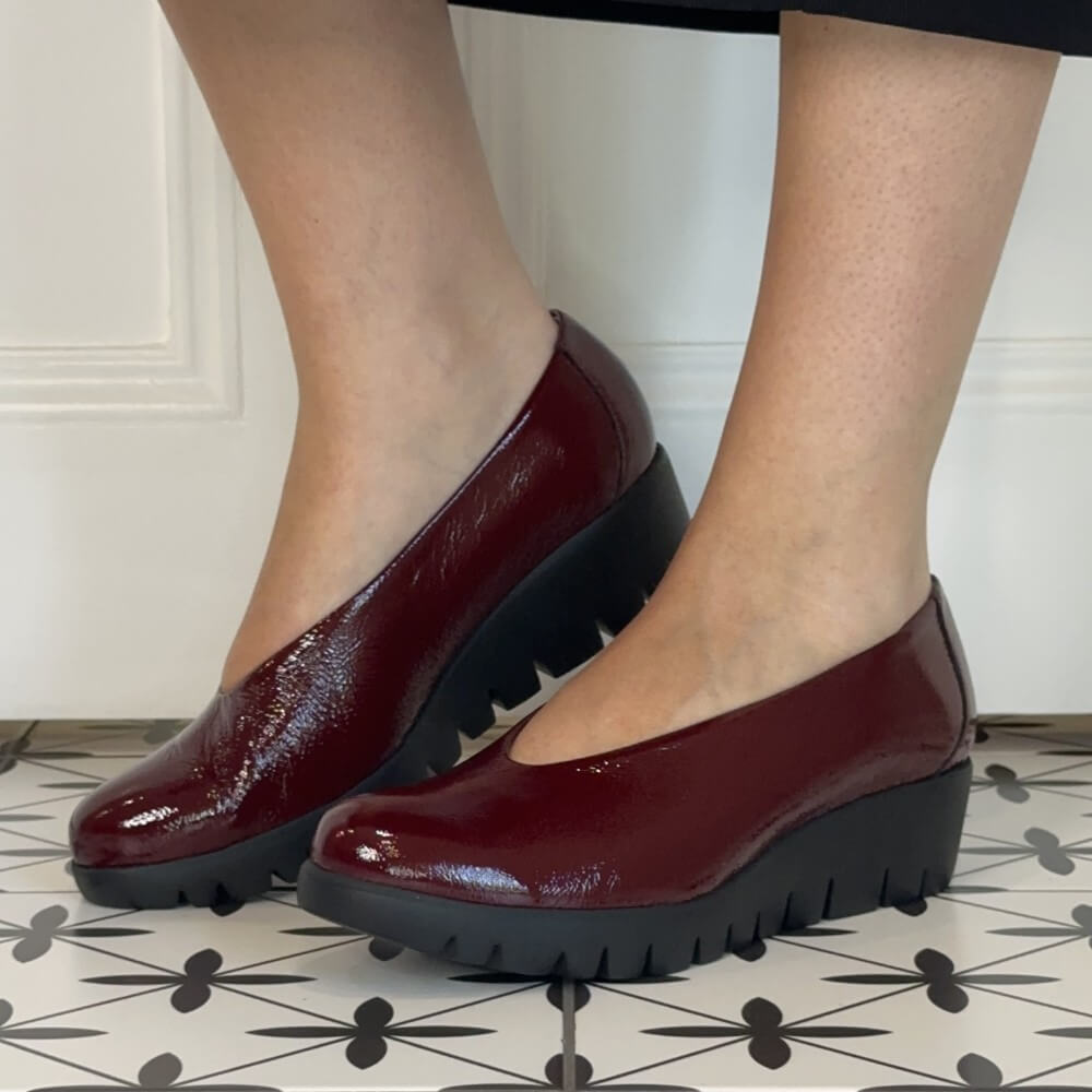 A pair of Wonders glossy burgundy platform wedge pumps with black lug soles, shown on a person's feet against a white panelled wall and patterned tile floor.