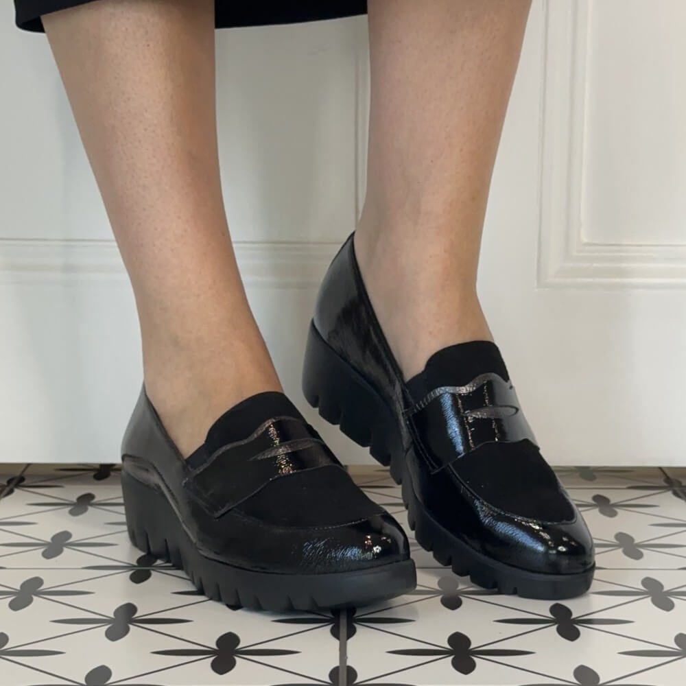 A person wearing Wonders black platform loafers with a glossy patent finish, shown from the knees down standing on a black-and-white patterned tile floor against a white panelled wall.