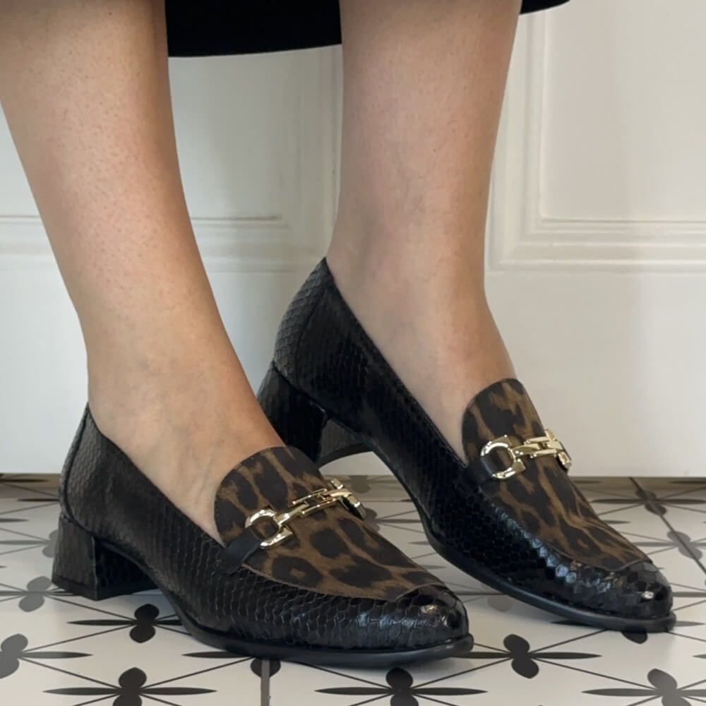 A person wearing wonders black snakeskin and leopard-print loafers with gold-tone horsebit details, shown from the knees down on a patterned tile floor.