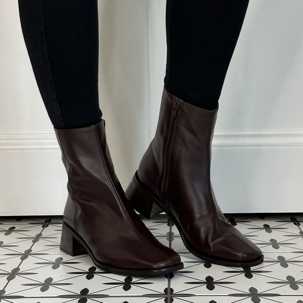 A pair of Wonders dark brown leather ankle boots with side zippers, worn with black leggings on a patterned black and white tile floor.