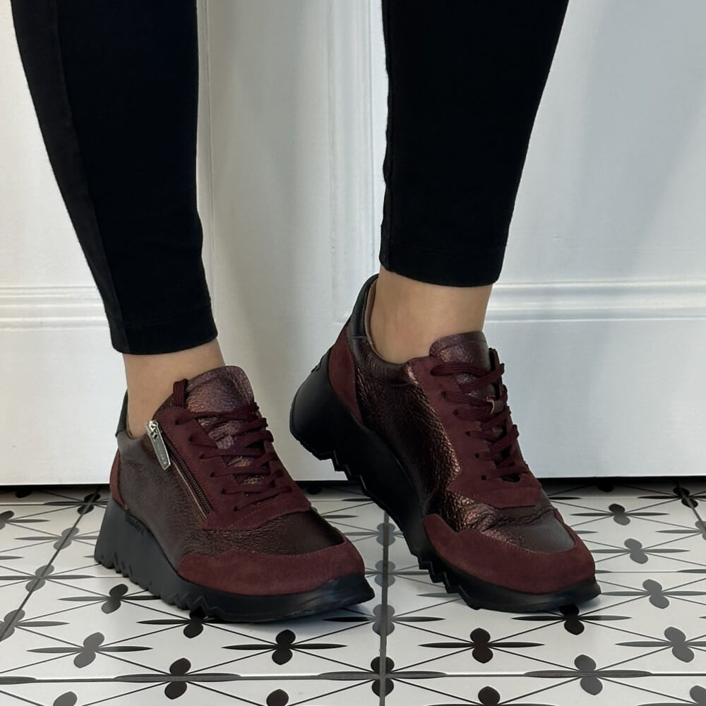 A person wearing Wonders burgundy and black platform sneakers with black leggings, standing on a black and white patterned tile floor in front of a white wall.