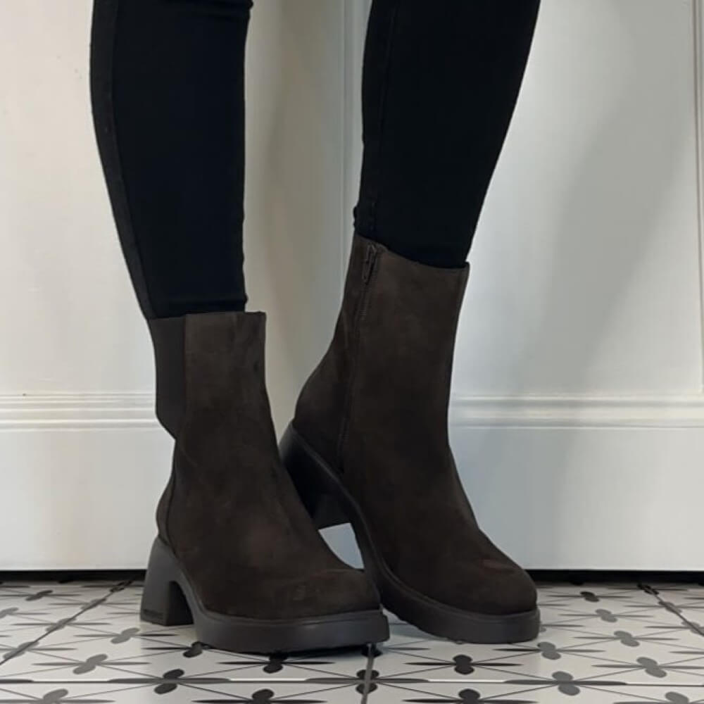 A pair of Wonders dark brown suede Chelsea-style ankle boots with elastic side panels and chunky black block heels, shown on a person wearing black leggings on a patterned floor.