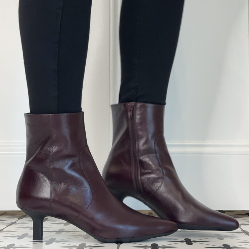 A pair of Wonders dark burgundy leather ankle boots with kitten heels, shown on a model wearing black leggings against a white wall and patterned tile floor.