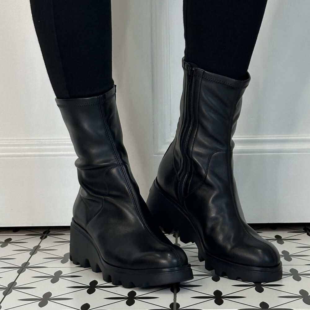 Wonders Black leather ankle boots with chunky wedge lug soles and side zippers, shown on a model wearing black leggings on a black and white patterned tile floor.