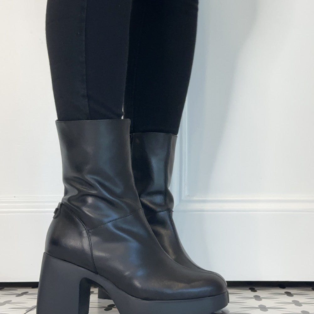 Wonders Black leather ankle boots with chunky block heels and platform soles, shown worn with black leggings against a white panelled wall.
