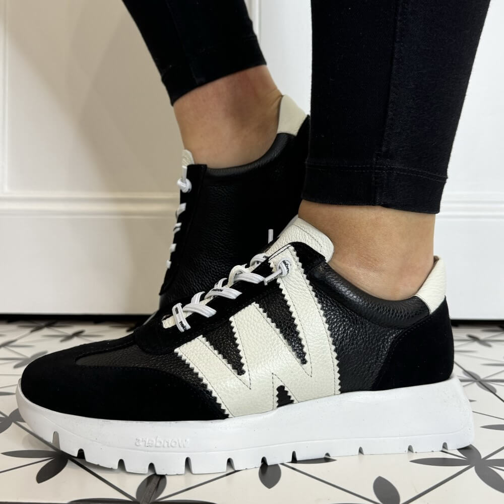 Black and white sneakers worn with black pants on a patterned floor.