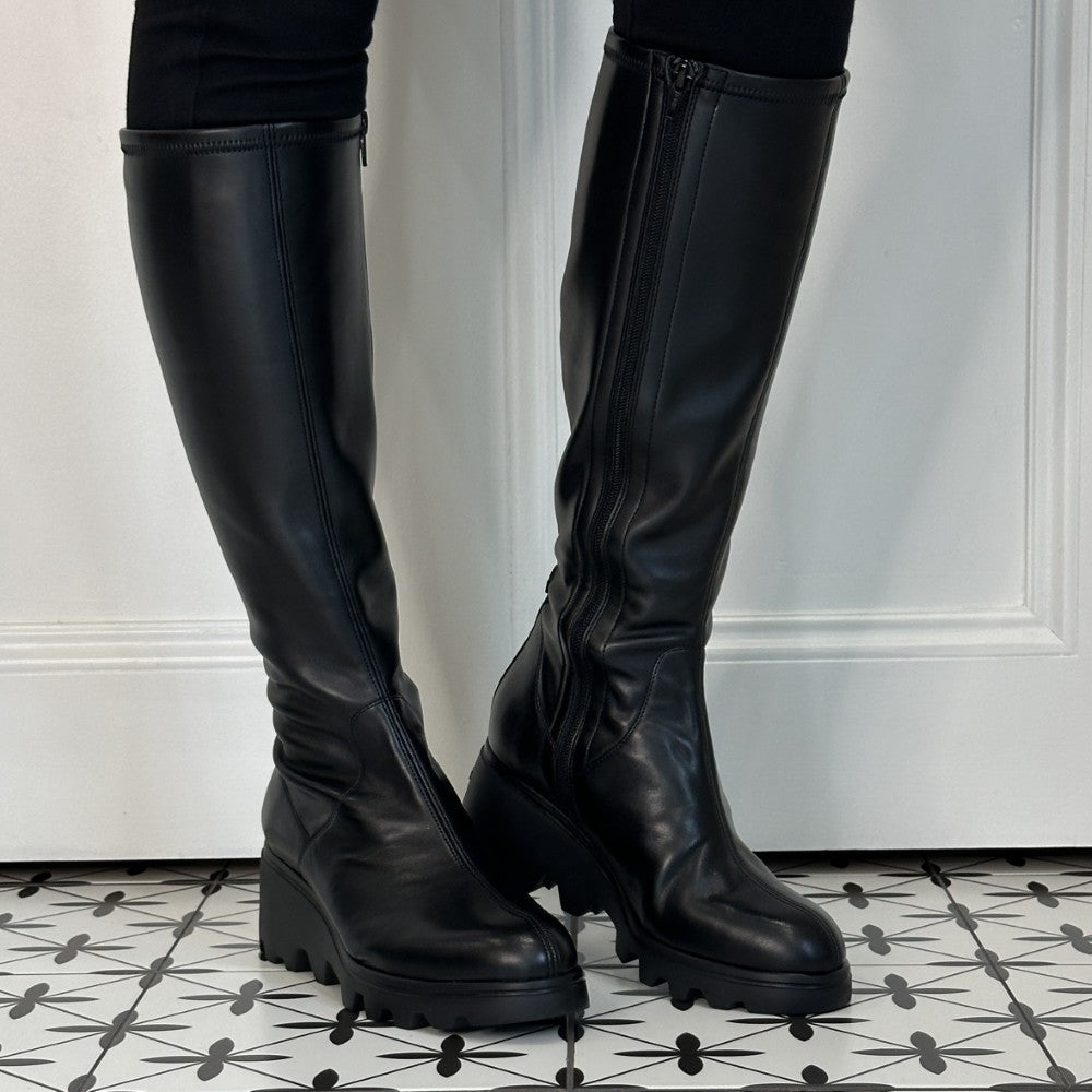 A pair of Wonders black leather knee-high boots with side zippers and chunky lug soles, shown worn with black leggings on a patterned black-and-white tile floor against a white panelled wall.