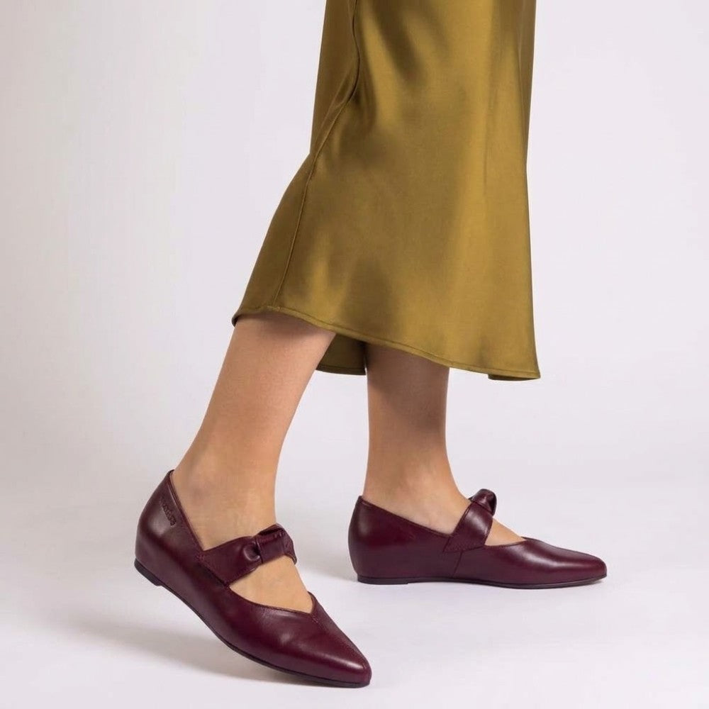 A person wearing Wonders dark burgundy leather Mary Jane flats with a bow detail, paired with an olive green satin skirt, shown from the knees down against a light gray studio background.