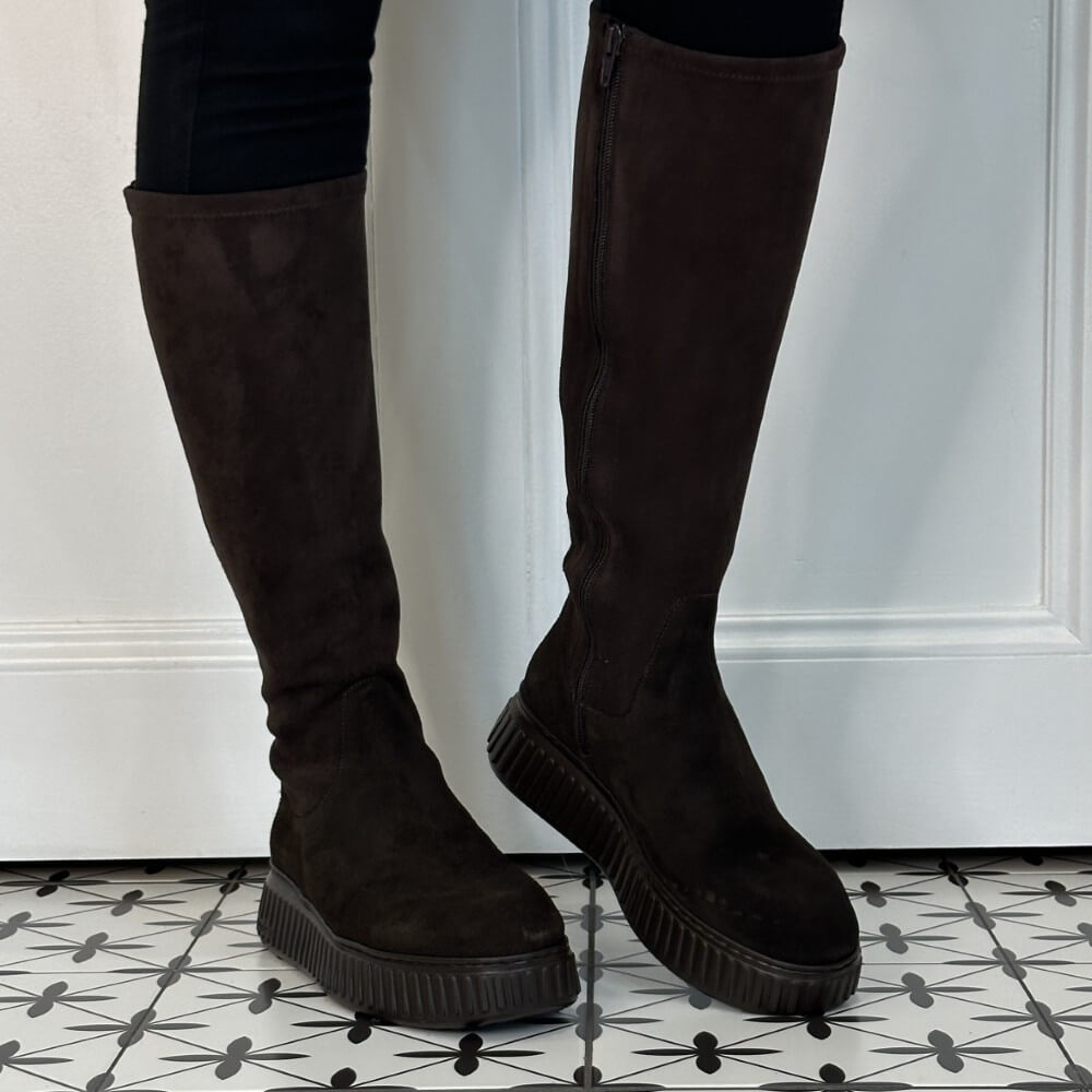 A pair of Wonders dark brown suede knee-high platform boots with side zippers, shown on a model's legs against a white panelled wall and black-and-white patterned tile floor.