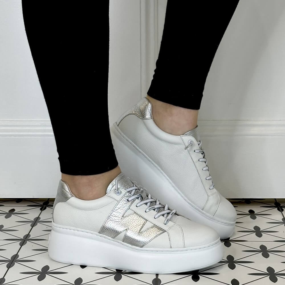 White sneakers with silver accents worn by a person on a patterned floor.