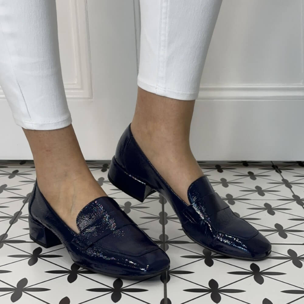 Navy blue patent loafers worn with white pants on a patterned floor.
