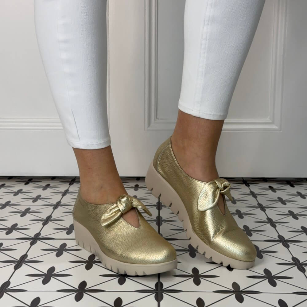 Gold shoes with bow details worn with white pants on a patterned floor.