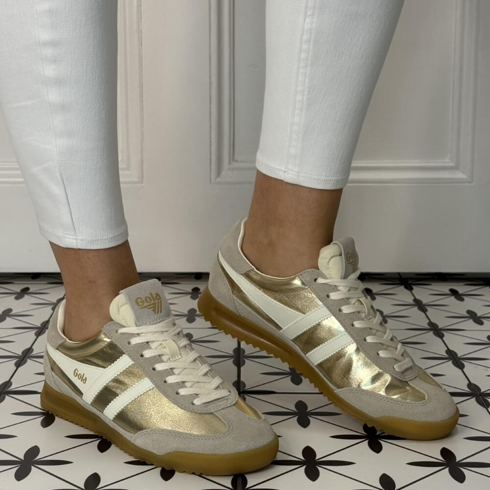 Gola Firefly Gold and beige sneakers worn with white pants on a patterned floor.