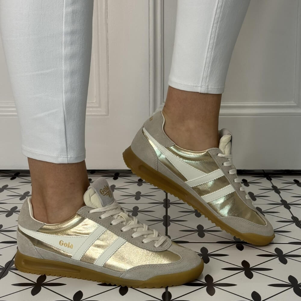 Gola Firefly Gold and beige sneakers worn with white pants on a patterned floor.