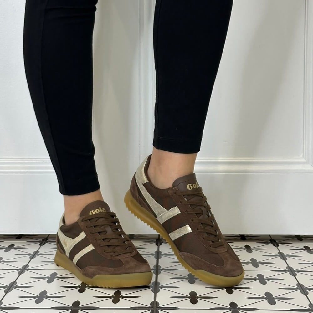 A person wearing Gola dark brown suede and leather sneakers with metallic gold side stripes, paired with black leggings, standing on a black-and-white patterned tile floor.