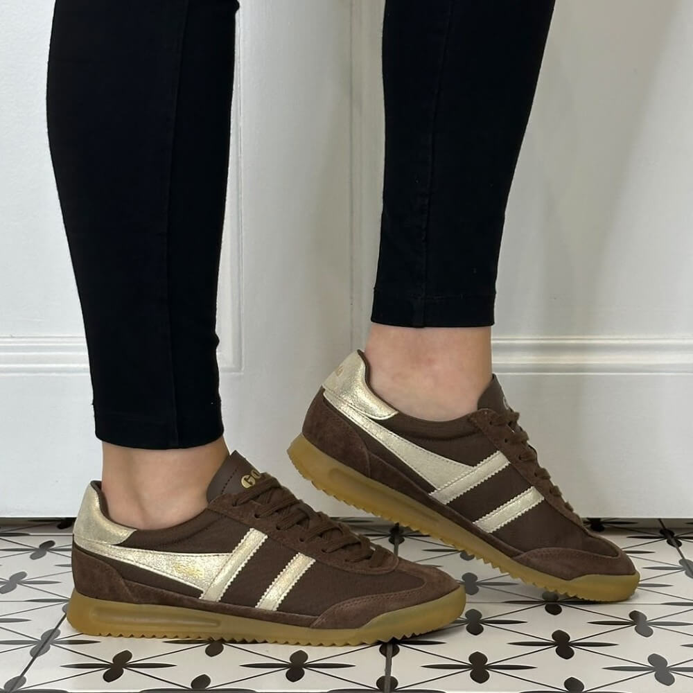 A person wearing Gola dark brown suede and leather sneakers with metallic gold side stripes, paired with black leggings, standing on a black-and-white patterned tile floor.